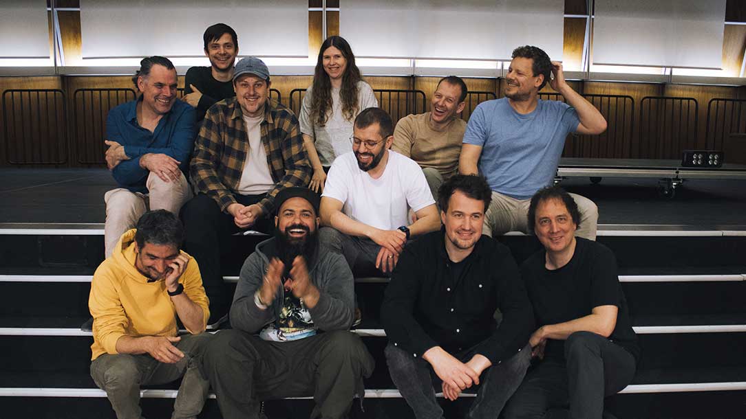 Gruppenporträt von elf Musikerinnen und Musikern, die in mehreren Reihen auf einer Tribüne sitzen und in ungezwungener, fröhlicher Atmosphäre miteinander lachen.
